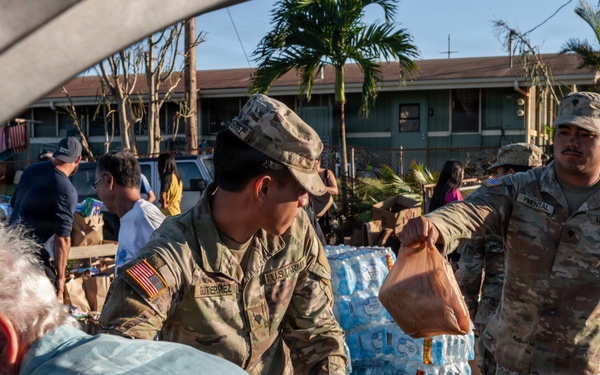 U.S. Army soldiers are distributing relief supplies, including bottled water and food bags, to civilians at what appears to be a disaster relief distribution point. Multiple soldiers in OCP (Operational Camouflage Pattern) uniforms with U.S. Army patches are actively handing out supplies. Large pallets of bottled water are visible in the background, suggesting a organized emergency relief operation.