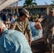 U.S. Army soldiers are distributing relief supplies, including bottled water and food bags, to civilians at what appears to be a disaster relief distribution point. Multiple soldiers in OCP (Operational Camouflage Pattern) uniforms with U.S. Army patches are actively handing out supplies. Large pallets of bottled water are visible in the background, suggesting a organized emergency relief operation.