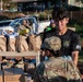 Members of the Hawaii Army National Guard are conducting a food distribution operation outdoors. Personnel in military camouflage uniforms and a civilian volunteer in a Hawaii Army National Guard t-shirt are organizing and distributing pre-packed brown paper bags filled with food items. Cardboard boxes labeled 'SDR-F' with meal count information are visible in the foreground.