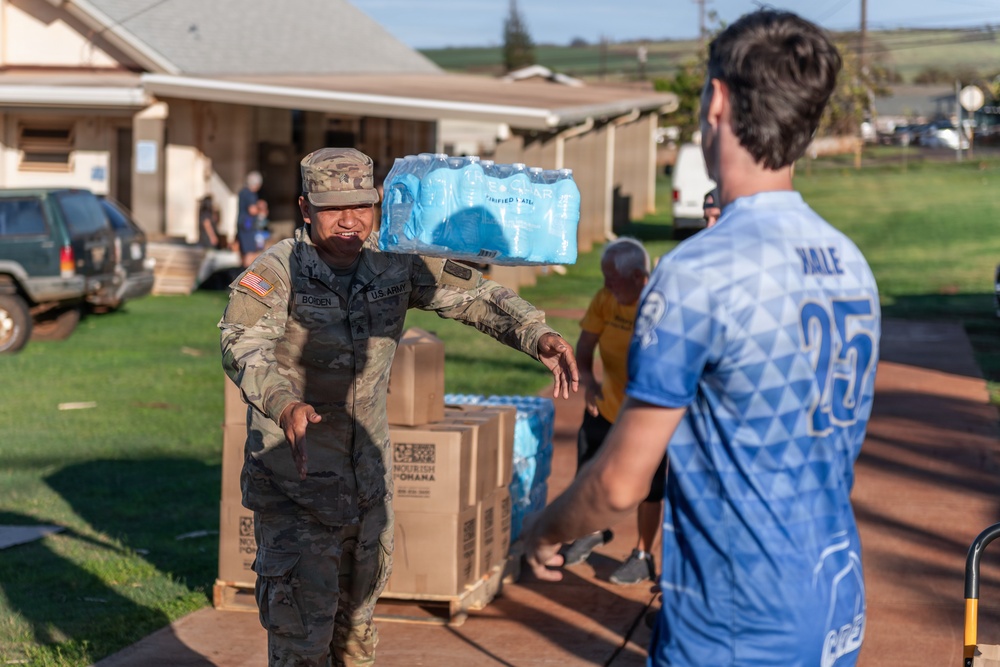 A U.S. Army soldier identified by name tape as 'BOLDEN' carries a case of bottled water on their shoulder while handing off supplies to a civilian wearing a blue sports jersey numbered 25. The scene appears to be a disaster relief or humanitarian aid distribution effort, with pallets of boxed goods and water visible in the background. The setting is an outdoor area near residential structures, likely in Hawaii based on the landscape.
