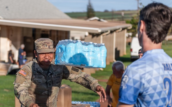 A U.S. Army soldier identified by name tape as 'BOLDEN' carries a case of bottled water on their shoulder while handing off supplies to a civilian wearing a blue sports jersey numbered 25. The scene appears to be a disaster relief or humanitarian aid distribution effort, with pallets of boxed goods and water visible in the background. The setting is an outdoor area near residential structures, likely in Hawaii based on the landscape.