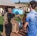 A U.S. Army soldier identified by name tape as 'BOLDEN' carries a case of bottled water on their shoulder while handing off supplies to a civilian wearing a blue sports jersey numbered 25. The scene appears to be a disaster relief or humanitarian aid distribution effort, with pallets of boxed goods and water visible in the background. The setting is an outdoor area near residential structures, likely in Hawaii based on the landscape.