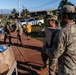 Military personnel in OCP (Operational Camouflage Pattern) uniforms are conducting a food and supply distribution operation in what appears to be a residential area. Soldiers are handling cardboard boxes and paper bags filled with food items, including a visible box of Cheerios cereal. The setting appears to be a disaster relief or community assistance operation, likely in Hawaii based on the tropical vegetation and red soil visible.