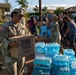 U.S. Army soldiers and civilians are conducting a disaster relief distribution operation, likely in Hawaii based on the 'Hawaii Food Bank - Nourish Our Ohana' box visible in the foreground. A soldier in Army Combat Uniform is carrying a food bank box while multiple cases of bottled water are stacked nearby. The scene suggests an emergency response effort in a residential neighborhood.