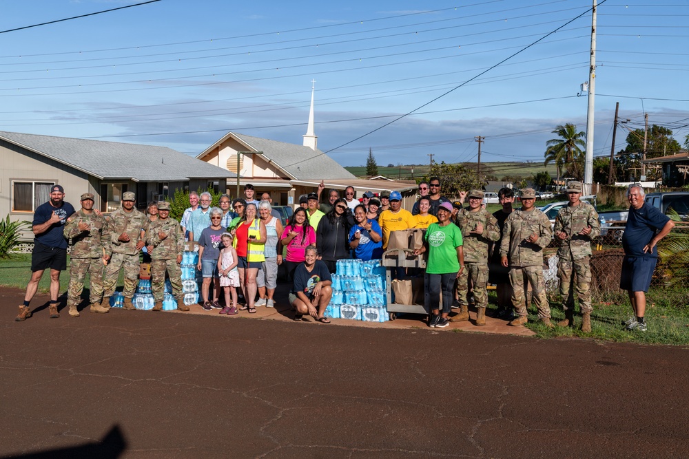 A large group of approximately 30 people, including military personnel in camouflage uniforms and civilians, pose together in front of a church building with a white steeple. Multiple cases of bottled water and paper bags are stacked in front of the group, suggesting a community relief or aid distribution effort. The setting appears to be a small town or rural community, likely in Hawaii based on the landscape and red soil.