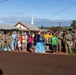 A large group of approximately 30 people, including military personnel in camouflage uniforms and civilians, pose together in front of a church building with a white steeple. Multiple cases of bottled water and paper bags are stacked in front of the group, suggesting a community relief or aid distribution effort. The setting appears to be a small town or rural community, likely in Hawaii based on the landscape and red soil.