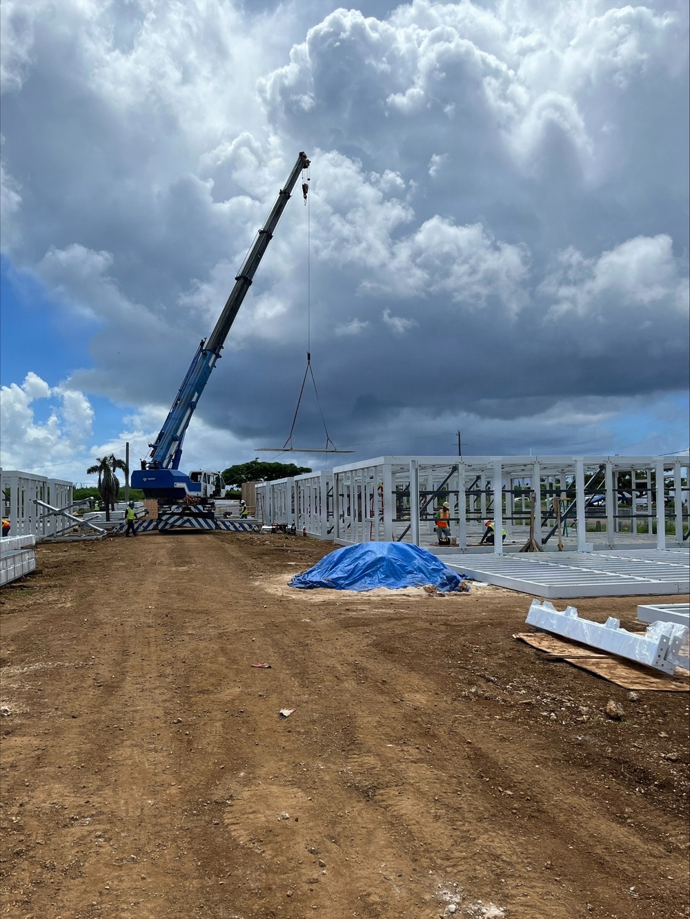 An active construction site showing the erection of a prefabricated steel or aluminum frame structure. A blue mobile crane is lifting a large panel or beam into position while workers in high-visibility vests oversee the assembly. The site features reddish-brown compacted dirt access roads, partially assembled wall frames, and dramatic cumulus cloud formations overhead.