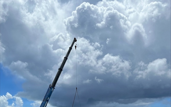 An active construction site showing the erection of a prefabricated steel or aluminum frame structure. A blue mobile crane is lifting a large panel or beam into position while workers in high-visibility vests oversee the assembly. The site features reddish-brown compacted dirt access roads, partially assembled wall frames, and dramatic cumulus cloud formations overhead.