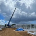An active construction site showing the erection of a prefabricated steel or aluminum frame structure. A blue mobile crane is lifting a large panel or beam into position while workers in high-visibility vests oversee the assembly. The site features reddish-brown compacted dirt access roads, partially assembled wall frames, and dramatic cumulus cloud formations overhead.