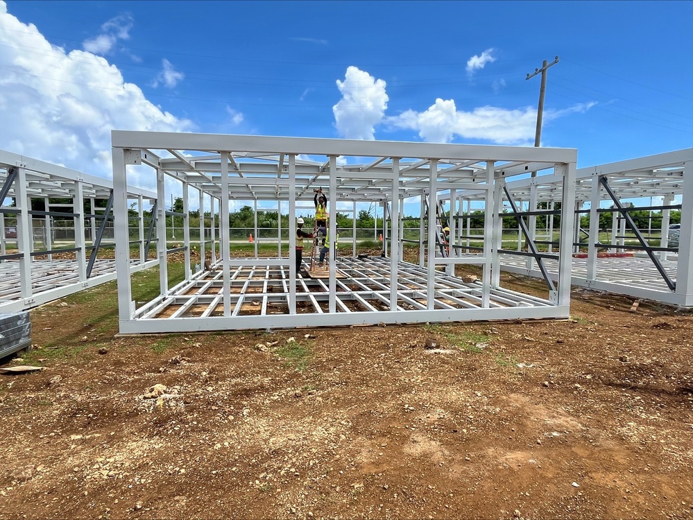 Construction workers are assembling a prefabricated steel-frame structure on a cleared dirt lot. The white metal framing system appears to be a modular or prefabricated building unit, with workers actively connecting components at height using a ladder. Multiple similar frame sections are visible extending to the left and right, suggesting a larger multi-unit construction project.