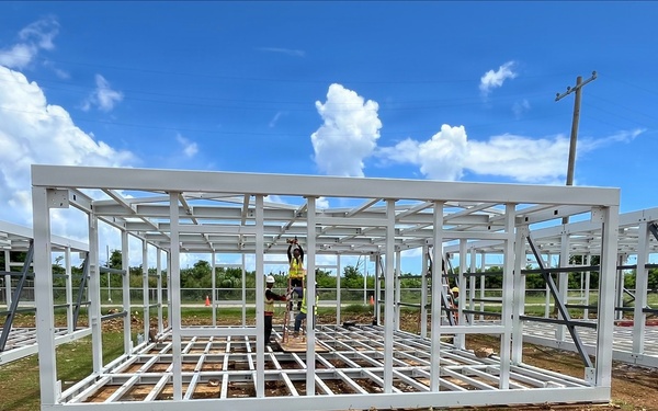 Construction workers are assembling a prefabricated steel-frame structure on a cleared dirt lot. The white metal framing system appears to be a modular or prefabricated building unit, with workers actively connecting components at height using a ladder. Multiple similar frame sections are visible extending to the left and right, suggesting a larger multi-unit construction project.