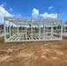 Construction workers are assembling a prefabricated steel-frame structure on a cleared dirt lot. The white metal framing system appears to be a modular or prefabricated building unit, with workers actively connecting components at height using a ladder. Multiple similar frame sections are visible extending to the left and right, suggesting a larger multi-unit construction project.