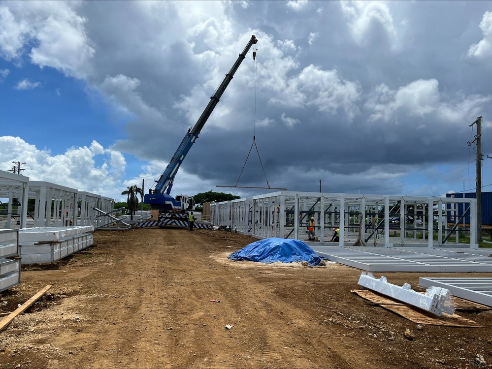 An active construction site showing the erection of a large prefabricated steel-frame structure. A blue mobile crane is lifting a steel beam or panel into position, with multiple workers visible on site. The setting appears to be a tropical or subtropical location based on the vegetation and sky conditions.
