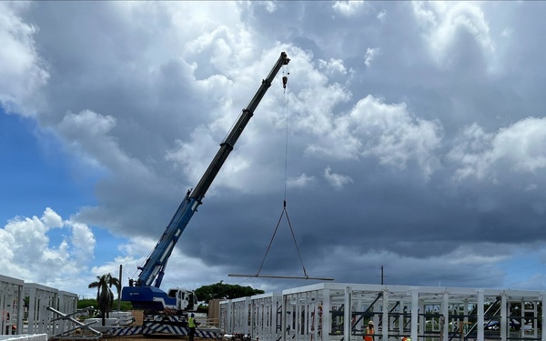 An active construction site showing the erection of a large prefabricated steel-frame structure. A blue mobile crane is lifting a steel beam or panel into position, with multiple workers visible on site. The setting appears to be a tropical or subtropical location based on the vegetation and sky conditions.