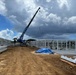 An active construction site showing the erection of a large prefabricated steel-frame structure. A blue mobile crane is lifting a steel beam or panel into position, with multiple workers visible on site. The setting appears to be a tropical or subtropical location based on the vegetation and sky conditions.