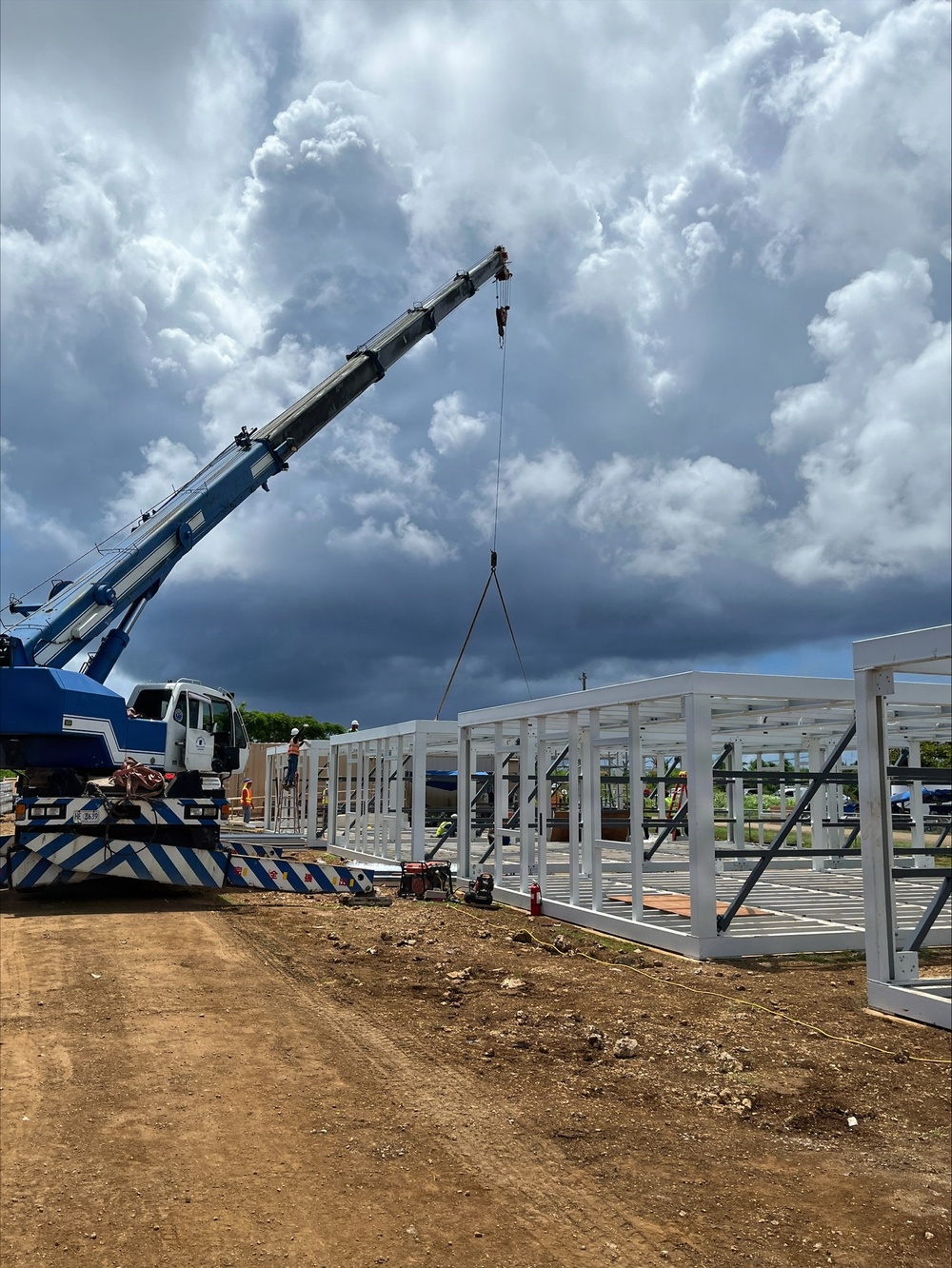 An active construction site showing the erection of a white steel-frame structure using a blue mobile telescopic crane. Multiple workers in high-visibility safety gear are visible on and around the partially assembled framework. The scene is set on a cleared dirt lot under dramatic, heavily clouded skies.