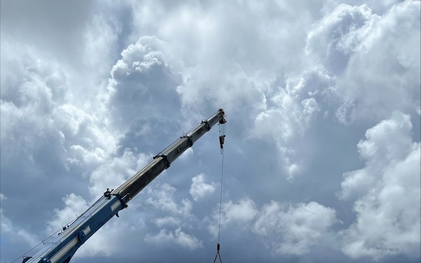 An active construction site showing the erection of a white steel-frame structure using a blue mobile telescopic crane. Multiple workers in high-visibility safety gear are visible on and around the partially assembled framework. The scene is set on a cleared dirt lot under dramatic, heavily clouded skies.