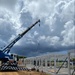 An active construction site showing the erection of a white steel-frame structure using a blue mobile telescopic crane. Multiple workers in high-visibility safety gear are visible on and around the partially assembled framework. The scene is set on a cleared dirt lot under dramatic, heavily clouded skies.