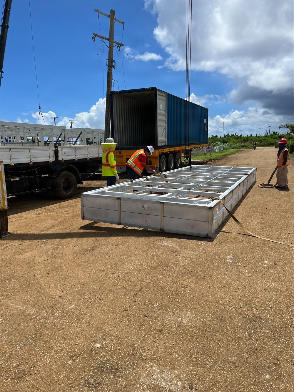 A construction or logistics operation is underway at an outdoor dirt lot, where workers in high-visibility safety gear are unloading or positioning a large prefabricated metal frame structure from a shipping container on a flatbed trailer. The metal frame, which appears to be a modular panel or structural component with visible labeling (XD1-2, XD1-3), is lying flat on the ground with crane cables attached. The setting appears to be a rural or semi-developed area with utility poles, sparse vegetation, and a secondary flatbed truck visible to the left.
