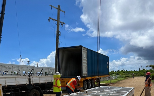 A construction or logistics operation is underway at an outdoor dirt lot, where workers in high-visibility safety gear are unloading or positioning a large prefabricated metal frame structure from a shipping container on a flatbed trailer. The metal frame, which appears to be a modular panel or structural component with visible labeling (XD1-2, XD1-3), is lying flat on the ground with crane cables attached. The setting appears to be a rural or semi-developed area with utility poles, sparse vegetation, and a secondary flatbed truck visible to the left.