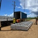 A construction or logistics operation is underway at an outdoor dirt lot, where workers in high-visibility safety gear are unloading or positioning a large prefabricated metal frame structure from a shipping container on a flatbed trailer. The metal frame, which appears to be a modular panel or structural component with visible labeling (XD1-2, XD1-3), is lying flat on the ground with crane cables attached. The setting appears to be a rural or semi-developed area with utility poles, sparse vegetation, and a secondary flatbed truck visible to the left.