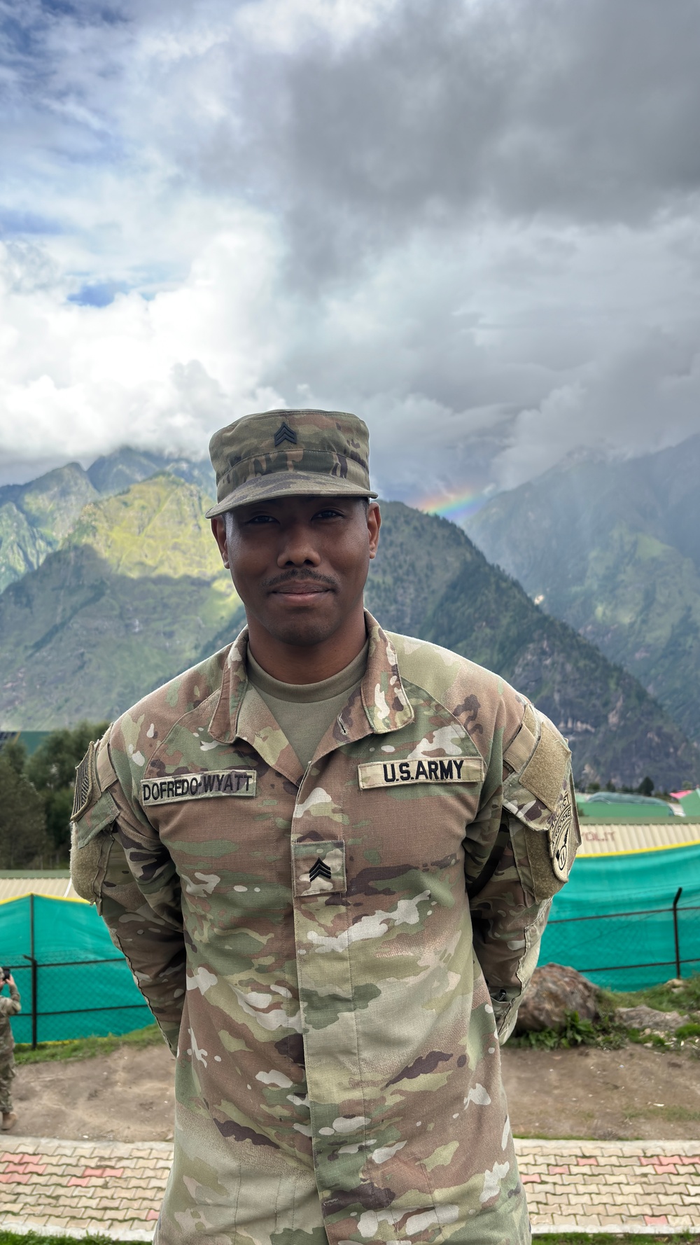A U.S. Army Sergeant named Dofredo-Wyatt is photographed in full MultiCam (OCP) uniform against a mountainous backdrop with dramatic cloud cover and a visible rainbow. The soldier stands at ease with hands behind their back in what appears to be a forward operating or training location. Green temporary structures and fencing are visible in the background, suggesting a field camp environment.