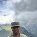 A U.S. Army Sergeant named Dofredo-Wyatt is photographed in full MultiCam (OCP) uniform against a mountainous backdrop with dramatic cloud cover and a visible rainbow. The soldier stands at ease with hands behind their back in what appears to be a forward operating or training location. Green temporary structures and fencing are visible in the background, suggesting a field camp environment.