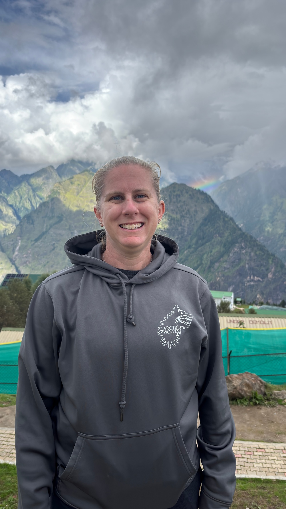 A person stands outdoors smiling at the camera, wearing a gray hoodie with an 'Arctic Wolves' logo on the chest. The background features dramatic mountain scenery with green slopes, storm clouds, and a faint rainbow visible. A green fence/netting and some structures are visible in the mid-ground.