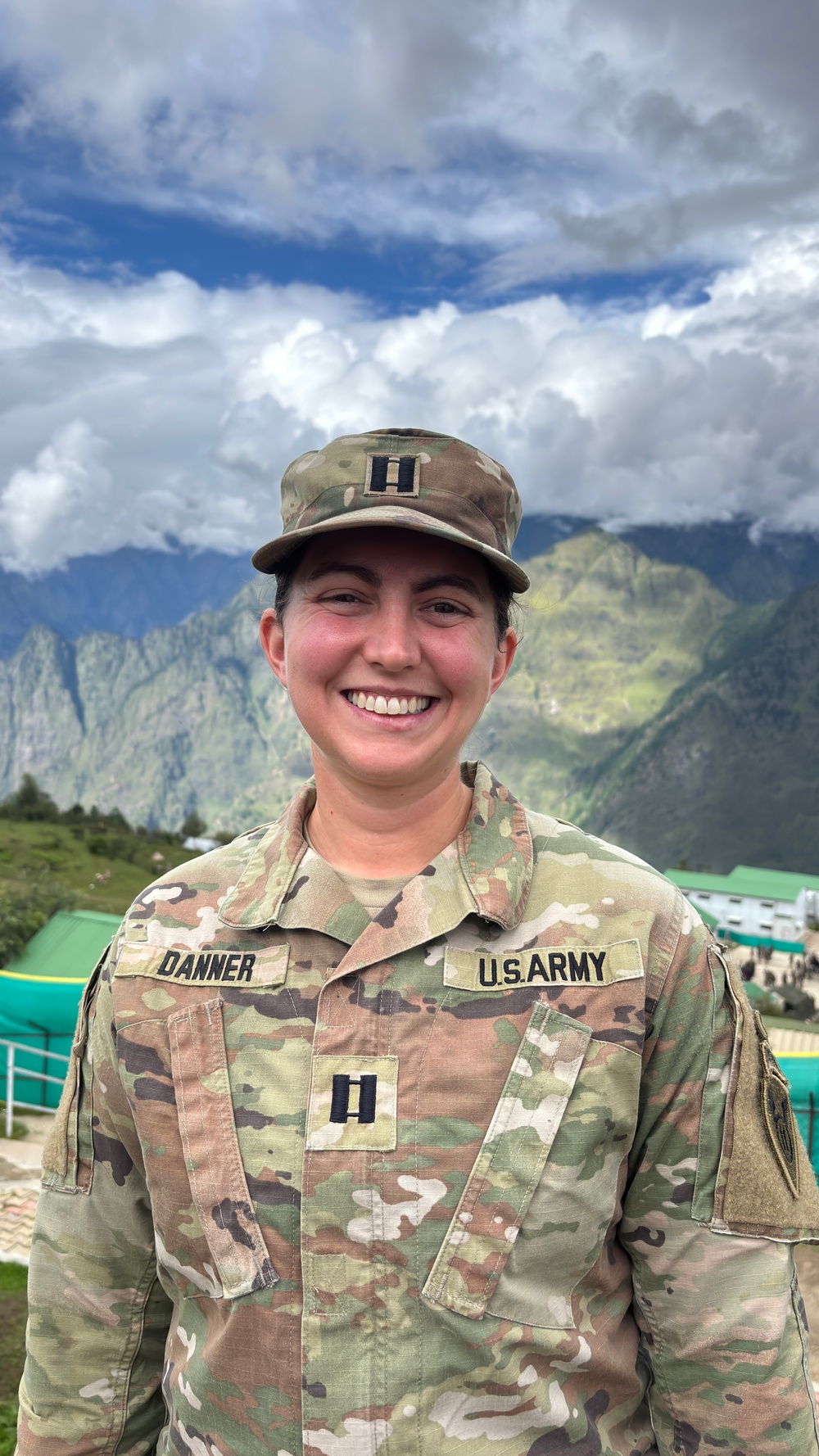 A U.S. Army Captain named Danner is photographed in uniform against a mountainous backdrop with dramatic cloud cover. She is smiling and wearing the Operational Camouflage Pattern (OCP) uniform with Captain rank insignia visible on both her patrol cap and chest. The setting appears to be a high-altitude location, possibly in a region such as South or Central Asia based on the terrain.
