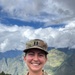 A U.S. Army Captain named Danner is photographed in uniform against a mountainous backdrop with dramatic cloud cover. She is smiling and wearing the Operational Camouflage Pattern (OCP) uniform with Captain rank insignia visible on both her patrol cap and chest. The setting appears to be a high-altitude location, possibly in a region such as South or Central Asia based on the terrain.