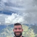 A smiling bearded man wearing a black 'ARMY' t-shirt with yellow lettering stands outdoors in front of a dramatic mountain landscape. The background features lush green mountains, large cumulus clouds, and what appears to be a sports facility or compound with green fencing. The man has visible tattoos on his left arm and appears to be in good spirits.