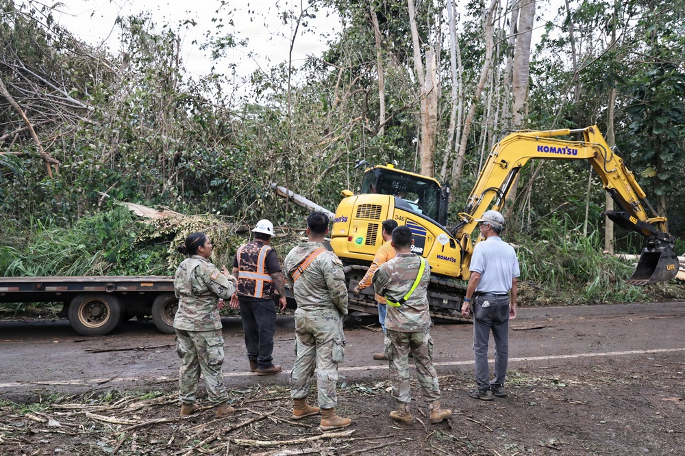 A group of military personnel in camouflage uniforms and civilian workers are gathered on a road surrounded by storm debris and downed trees. A yellow Komatsu excavator (model 346) is visible in the background, actively engaged in debris clearing operations. The scene appears to be post-storm disaster relief or road clearance work in a tropical or subtropical environment.