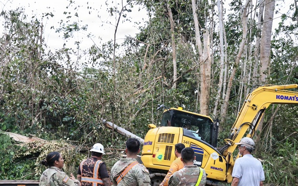 A group of military personnel in camouflage uniforms and civilian workers are gathered on a road surrounded by storm debris and downed trees. A yellow Komatsu excavator (model 346) is visible in the background, actively engaged in debris clearing operations. The scene appears to be post-storm disaster relief or road clearance work in a tropical or subtropical environment.