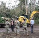 A group of military personnel in camouflage uniforms and civilian workers are gathered on a road surrounded by storm debris and downed trees. A yellow Komatsu excavator (model 346) is visible in the background, actively engaged in debris clearing operations. The scene appears to be post-storm disaster relief or road clearance work in a tropical or subtropical environment.