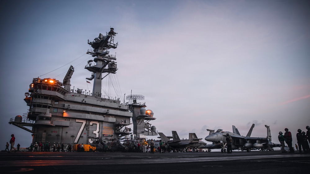 The image shows a U.S. Navy aircraft carrier flight deck, identifiable by the hull number '73' painted on the island superstructure, indicating USS George Washington (CVN-73). The scene appears to be taken at dusk or dawn, with multiple jet aircraft visible on the deck alongside numerous flight deck personnel. The carrier's island structure dominates the left side of the frame, featuring radar arrays, communication equipment, and navigation lights.