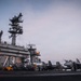 The image shows a U.S. Navy aircraft carrier flight deck, identifiable by the hull number '73' painted on the island superstructure, indicating USS George Washington (CVN-73). The scene appears to be taken at dusk or dawn, with multiple jet aircraft visible on the deck alongside numerous flight deck personnel. The carrier's island structure dominates the left side of the frame, featuring radar arrays, communication equipment, and navigation lights.