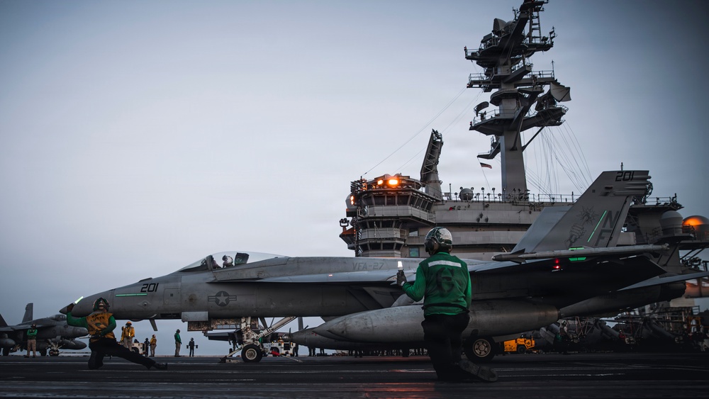 A U.S. Navy F/A-18 Super Hornet (Bureau Number 201, marked 'VFA-27') is positioned on the flight deck of an aircraft carrier, likely preparing for launch. Deck crew in green jerseys are performing pre-launch checks and signaling procedures. The carrier's island superstructure is visible in the background with illuminated lights, suggesting low-light or dusk conditions.