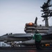 A U.S. Navy F/A-18 Super Hornet (Bureau Number 201, marked 'VFA-27') is positioned on the flight deck of an aircraft carrier, likely preparing for launch. Deck crew in green jerseys are performing pre-launch checks and signaling procedures. The carrier's island superstructure is visible in the background with illuminated lights, suggesting low-light or dusk conditions.