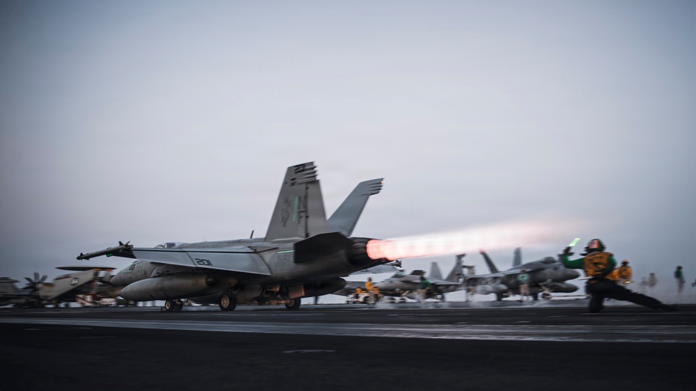 A carrier-based jet aircraft, appearing to be an F/A-18 Hornet or Super Hornet with modex number '201', is being catapult-launched from a naval aircraft carrier flight deck at dusk or dawn. The aircraft's engines are in full afterburner, producing a visible orange-white flame plume. Flight deck personnel in colored jerseys are visible in the background alongside additional parked aircraft.