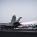 A carrier-based jet aircraft, appearing to be an F/A-18 Hornet or Super Hornet with modex number '201', is being catapult-launched from a naval aircraft carrier flight deck at dusk or dawn. The aircraft's engines are in full afterburner, producing a visible orange-white flame plume. Flight deck personnel in colored jerseys are visible in the background alongside additional parked aircraft.