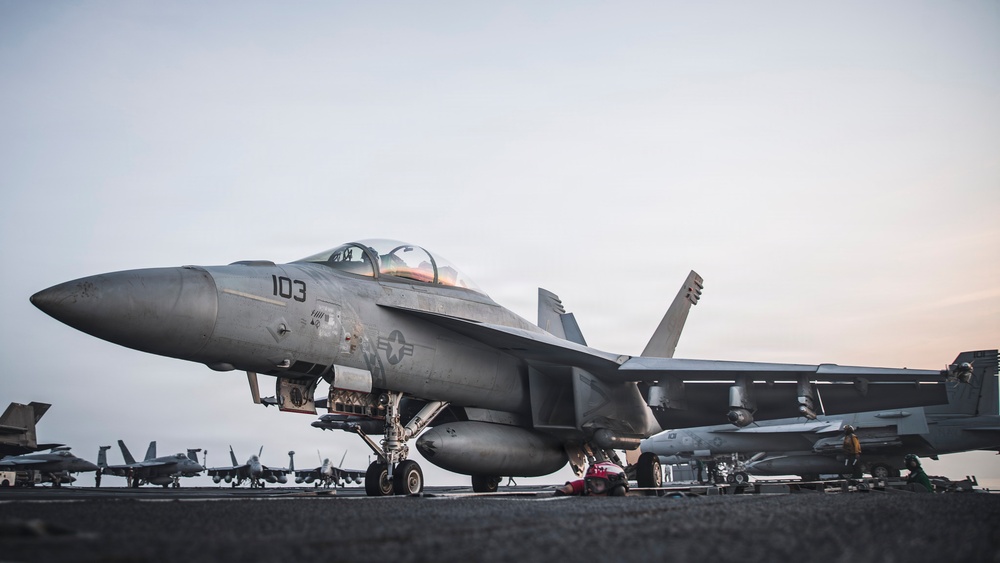 A U.S. Navy F/A-18 Super Hornet (modex 103) is positioned on the flight deck of an aircraft carrier, likely during dawn or dusk based on the overcast, muted sky. The aircraft is armed with visible ordnance and external fuel tanks, suggesting pre-mission or post-mission status. Multiple additional fighter aircraft are visible in the background, indicating a fully operational carrier air wing.