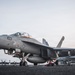 A U.S. Navy F/A-18 Super Hornet (modex 103) is positioned on the flight deck of an aircraft carrier, likely during dawn or dusk based on the overcast, muted sky. The aircraft is armed with visible ordnance and external fuel tanks, suggesting pre-mission or post-mission status. Multiple additional fighter aircraft are visible in the background, indicating a fully operational carrier air wing.