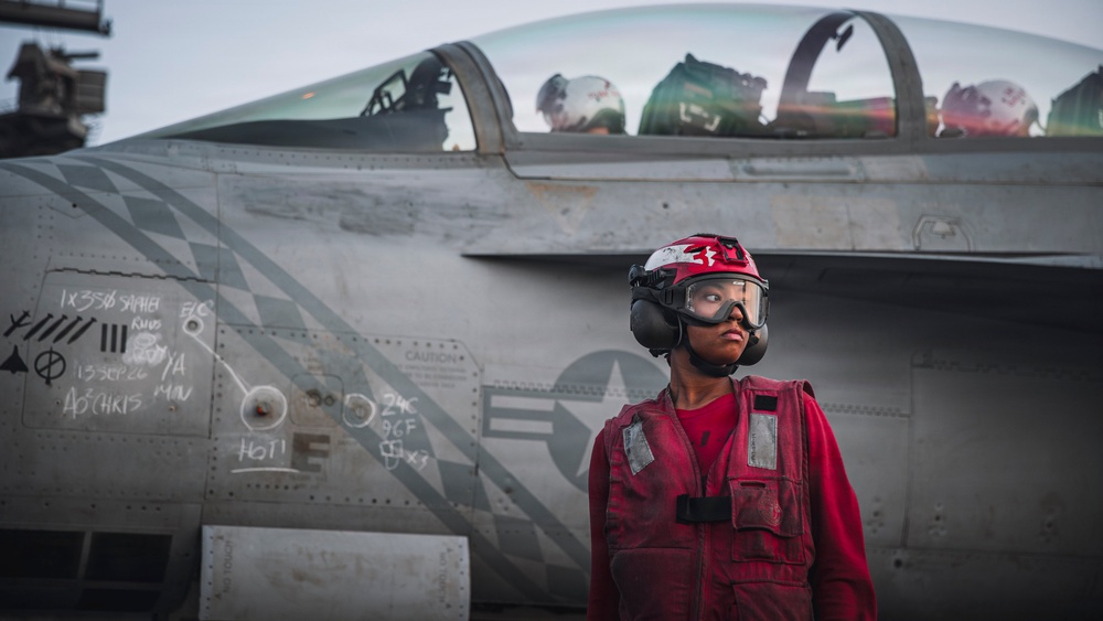 A U.S. Navy deck crew member in red gear stands in front of a carrier-based fighter jet, likely an F/A-18 Hornet or Super Hornet, on an aircraft carrier flight deck. The crew member is wearing standard red jersey, helmet, goggles, and hearing protection, indicating an ordnance or crash/salvage role. The aircraft fuselage shows chalk maintenance markings and a U.S. military star insignia.