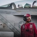 A U.S. Navy deck crew member in red gear stands in front of a carrier-based fighter jet, likely an F/A-18 Hornet or Super Hornet, on an aircraft carrier flight deck. The crew member is wearing standard red jersey, helmet, goggles, and hearing protection, indicating an ordnance or crash/salvage role. The aircraft fuselage shows chalk maintenance markings and a U.S. military star insignia.