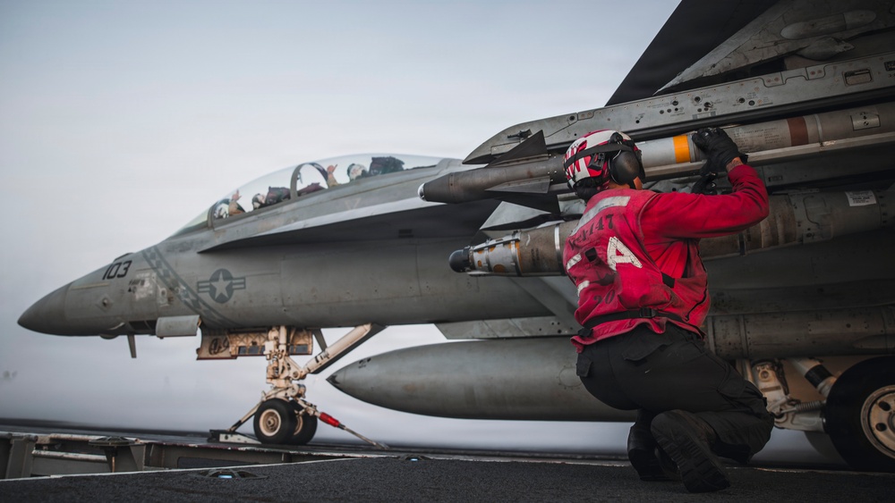 A U.S. Navy ordnance technician in a red jersey is performing weapons checks or maintenance on a carrier-based fighter jet on an aircraft carrier flight deck. The aircraft, marked with the number '103' and a U.S. military star insignia, appears to be an F/A-18 Super Hornet loaded with missiles and munitions. A pilot or crew member is visible in the cockpit in the background.