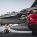 A U.S. Navy ordnance technician in a red jersey is performing weapons checks or maintenance on a carrier-based fighter jet on an aircraft carrier flight deck. The aircraft, marked with the number '103' and a U.S. military star insignia, appears to be an F/A-18 Super Hornet loaded with missiles and munitions. A pilot or crew member is visible in the cockpit in the background.
