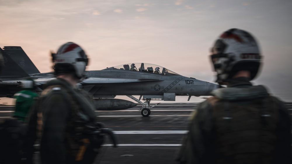 A U.S. Navy carrier-based jet aircraft, marked with the number '102' and a U.S. military star insignia, is positioned on an aircraft carrier flight deck at dusk or dawn. Two flight deck crew members wearing helmets and flight gear are visible in the foreground, out of focus. The scene depicts standard carrier flight operations with personnel observing or directing aircraft movement.