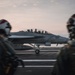 A U.S. Navy carrier-based jet aircraft, marked with the number '102' and a U.S. military star insignia, is positioned on an aircraft carrier flight deck at dusk or dawn. Two flight deck crew members wearing helmets and flight gear are visible in the foreground, out of focus. The scene depicts standard carrier flight operations with personnel observing or directing aircraft movement.