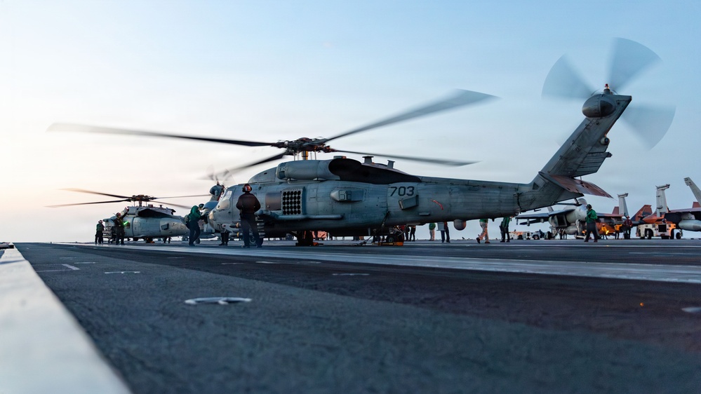 A military helicopter marked '703' with rotors spinning is positioned on the flight deck of a naval aircraft carrier, surrounded by deck crew in colored jerseys. At least two helicopters are visible, along with fixed-wing aircraft in the background. The scene appears to be captured during dawn or dusk based on the low ambient light conditions.