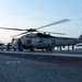 A military helicopter marked '703' with rotors spinning is positioned on the flight deck of a naval aircraft carrier, surrounded by deck crew in colored jerseys. At least two helicopters are visible, along with fixed-wing aircraft in the background. The scene appears to be captured during dawn or dusk based on the low ambient light conditions.